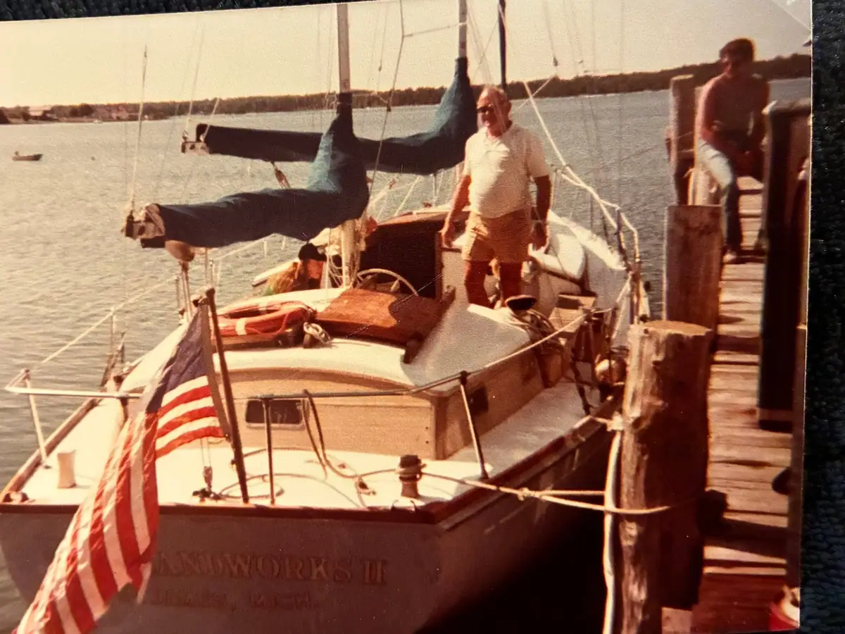 A sailboat with furled sails is moored beside a wooden pier, with two men nearby and an American flag at the bow.