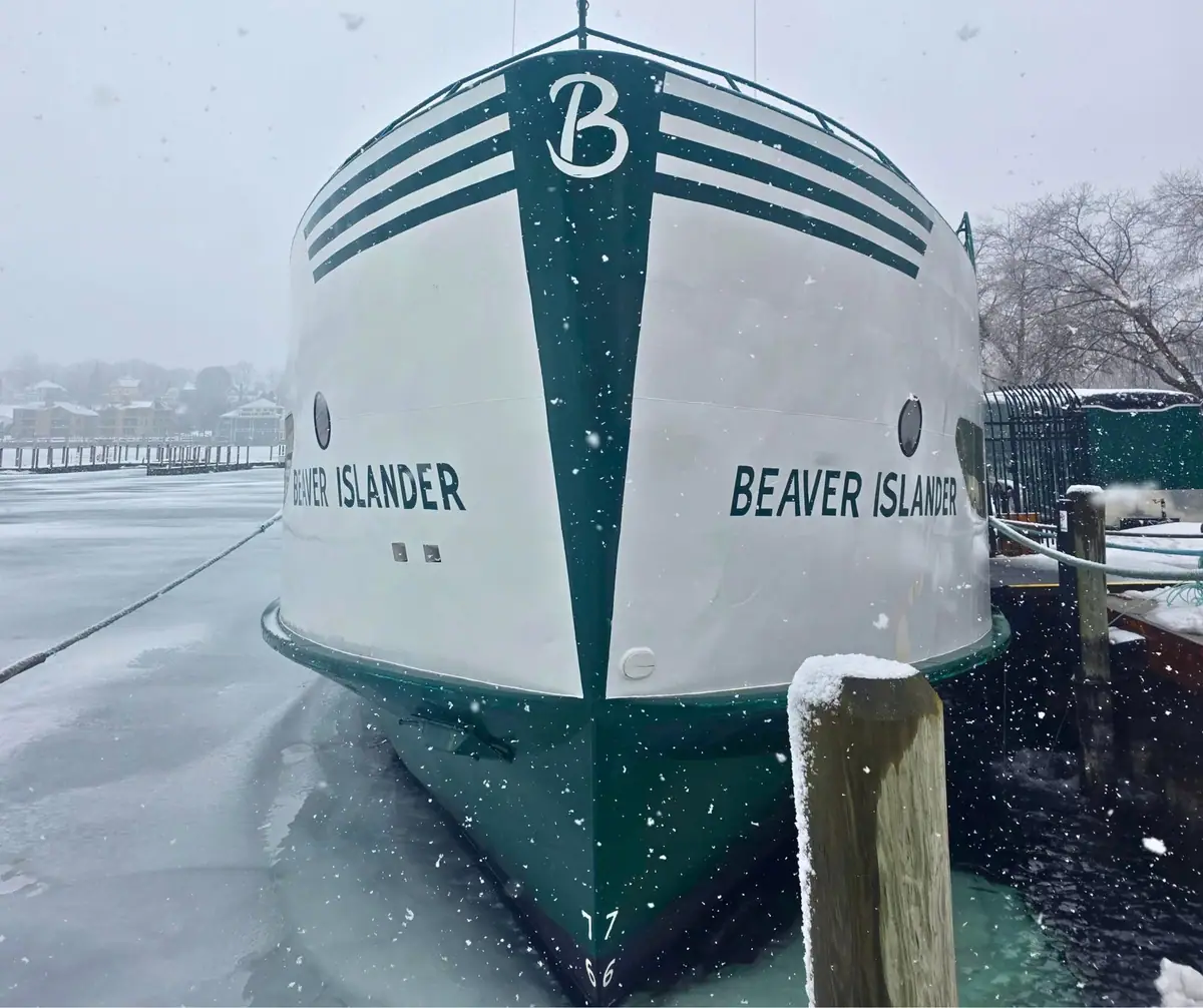 Snow-covered bow of the ferry BEAVER ISLANDER at a dock, with ice and a wooden piling in the foreground.