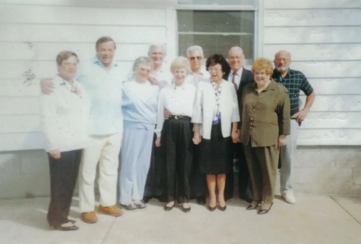 Ten adults pose together outdoors in front of a light-colored building with horizontal siding and a window.