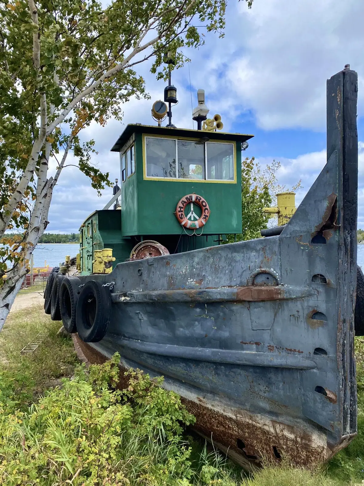 A weathered green tugboat with a peace-sign life ring stands beside the water beneath a cloudy sky.