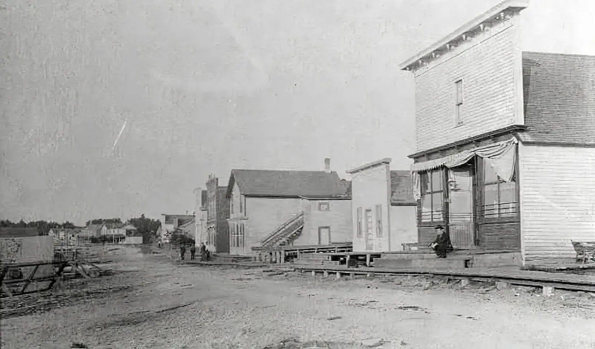 Black-and-white waterfront street scene with wooden commercial buildings, raised boardwalks, people, and fishing equipment.