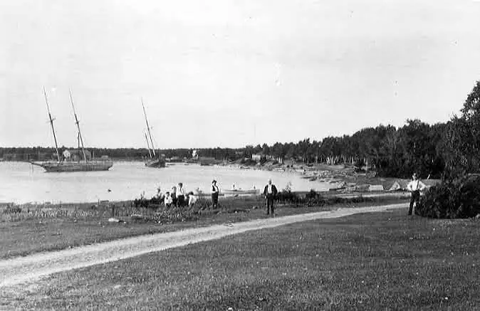 Black-and-white view of a curving shore road, people, trees, and sailing vessels in a sheltered bay.