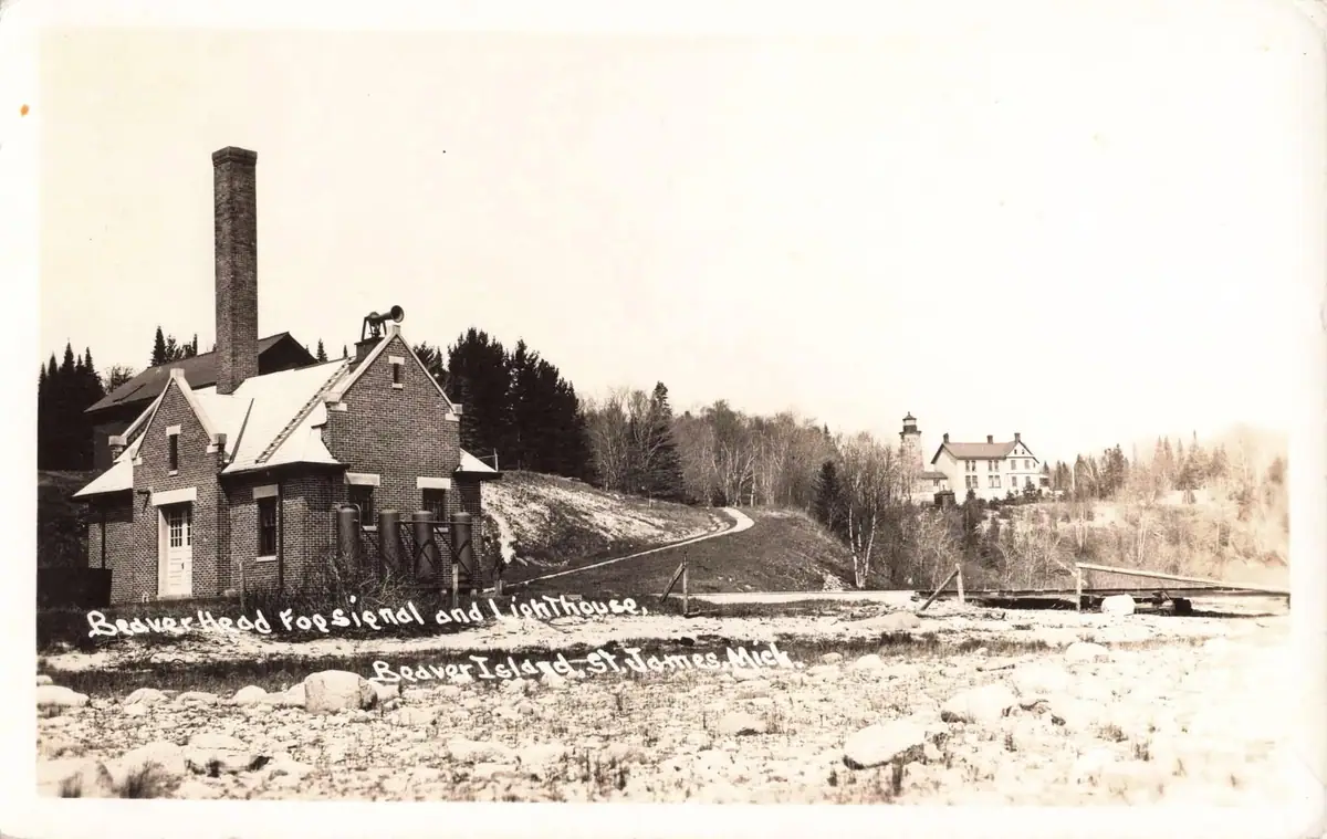 Brick fog-signal building and lighthouse beside a rocky shoreline, with a tree-covered hill behind.