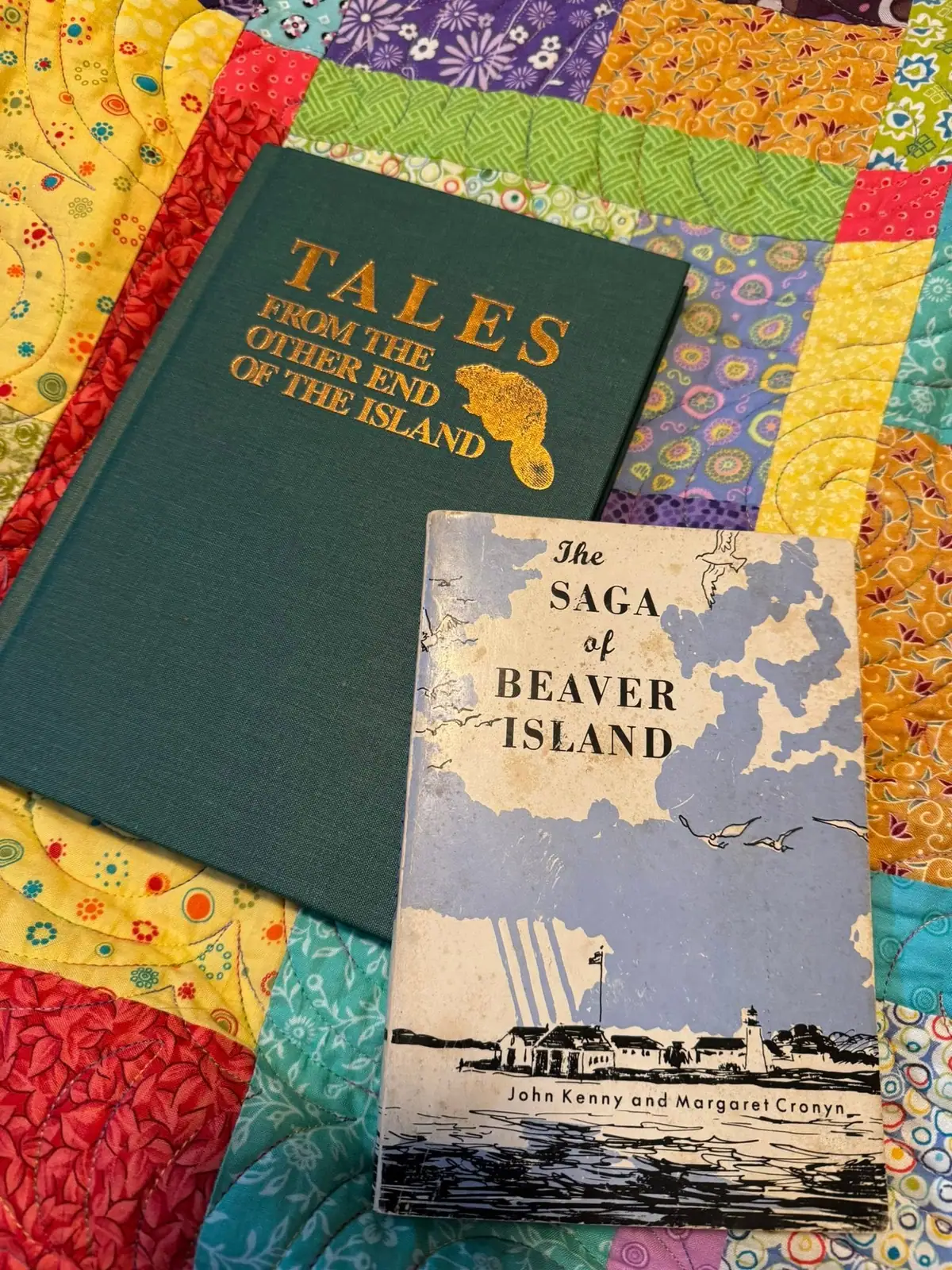 Two books rest on a colorful patchwork quilt, one showing an illustrated shoreline with a lighthouse.