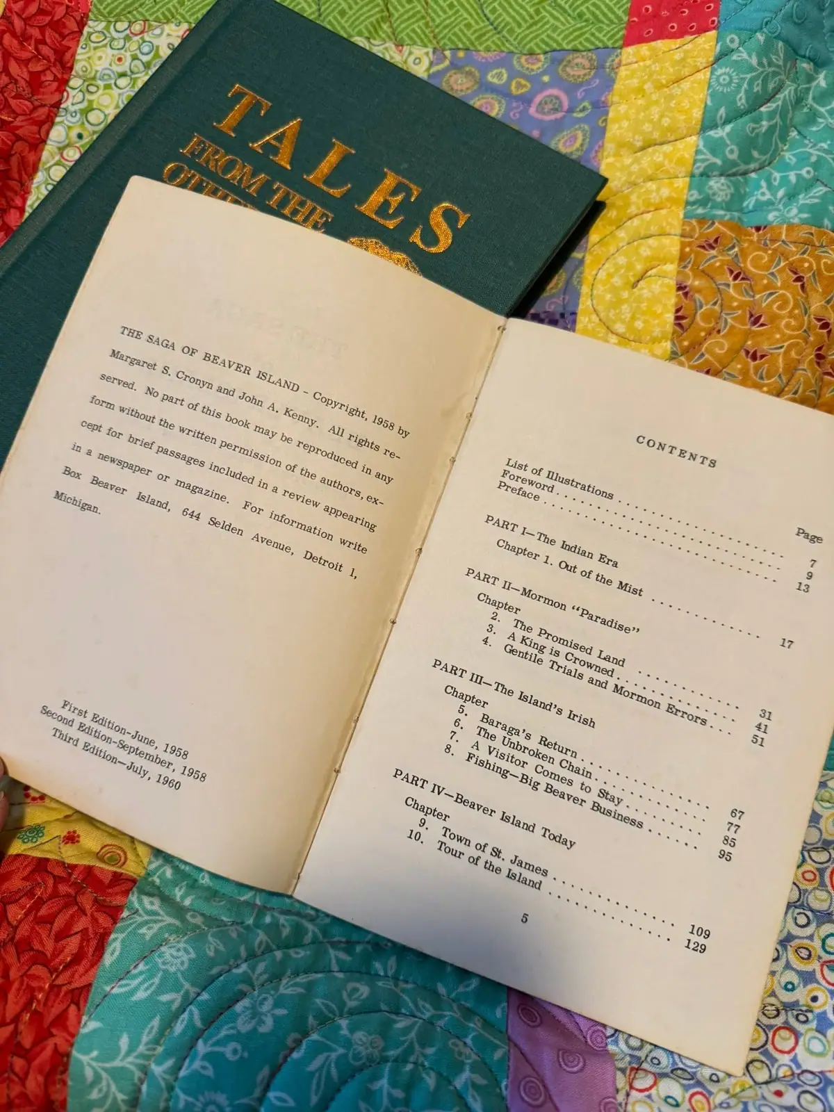 An open hardcover book with gold lettering rests on a colorful patchwork quilt.