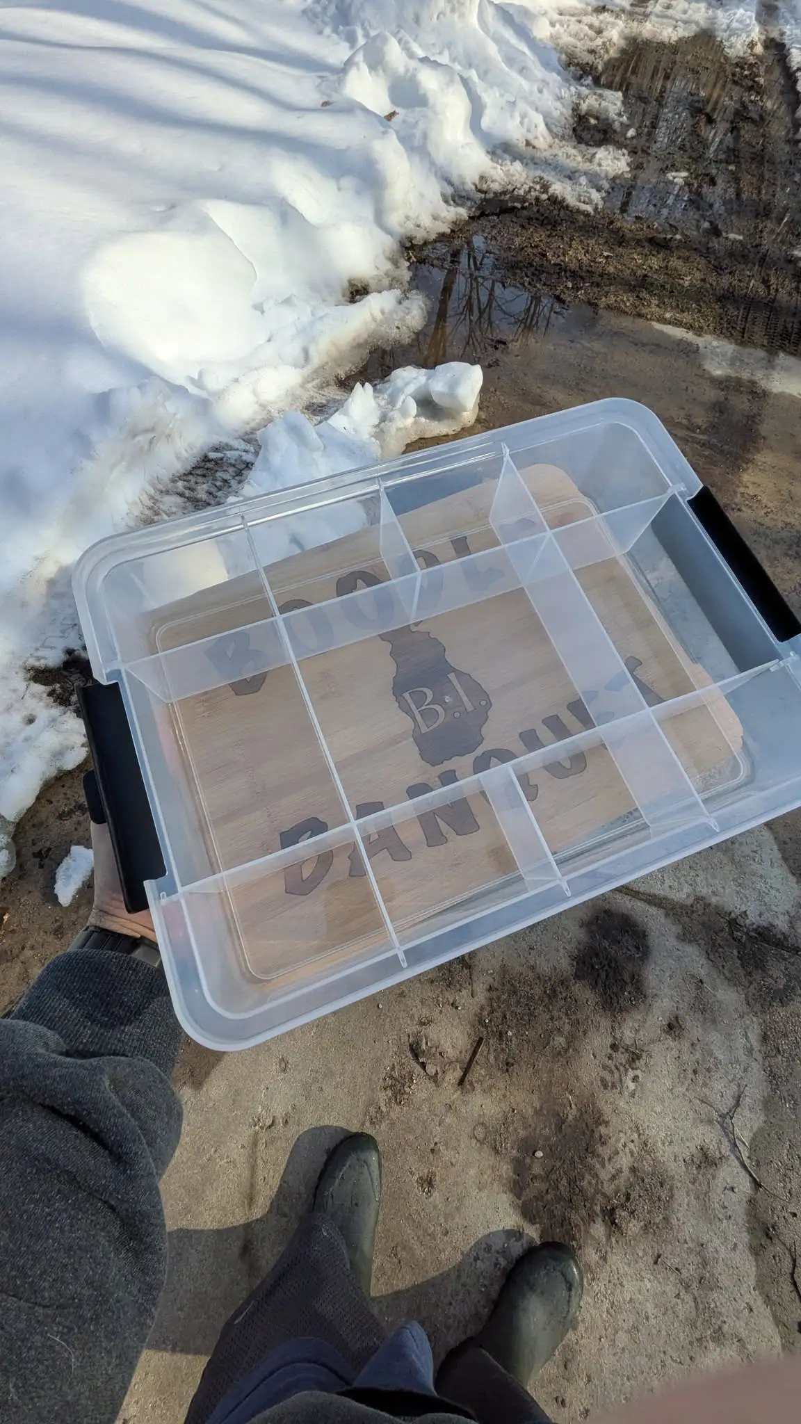 A clear compartmented storage box is held over snow and muddy ground, with lettering visible beneath its lid.