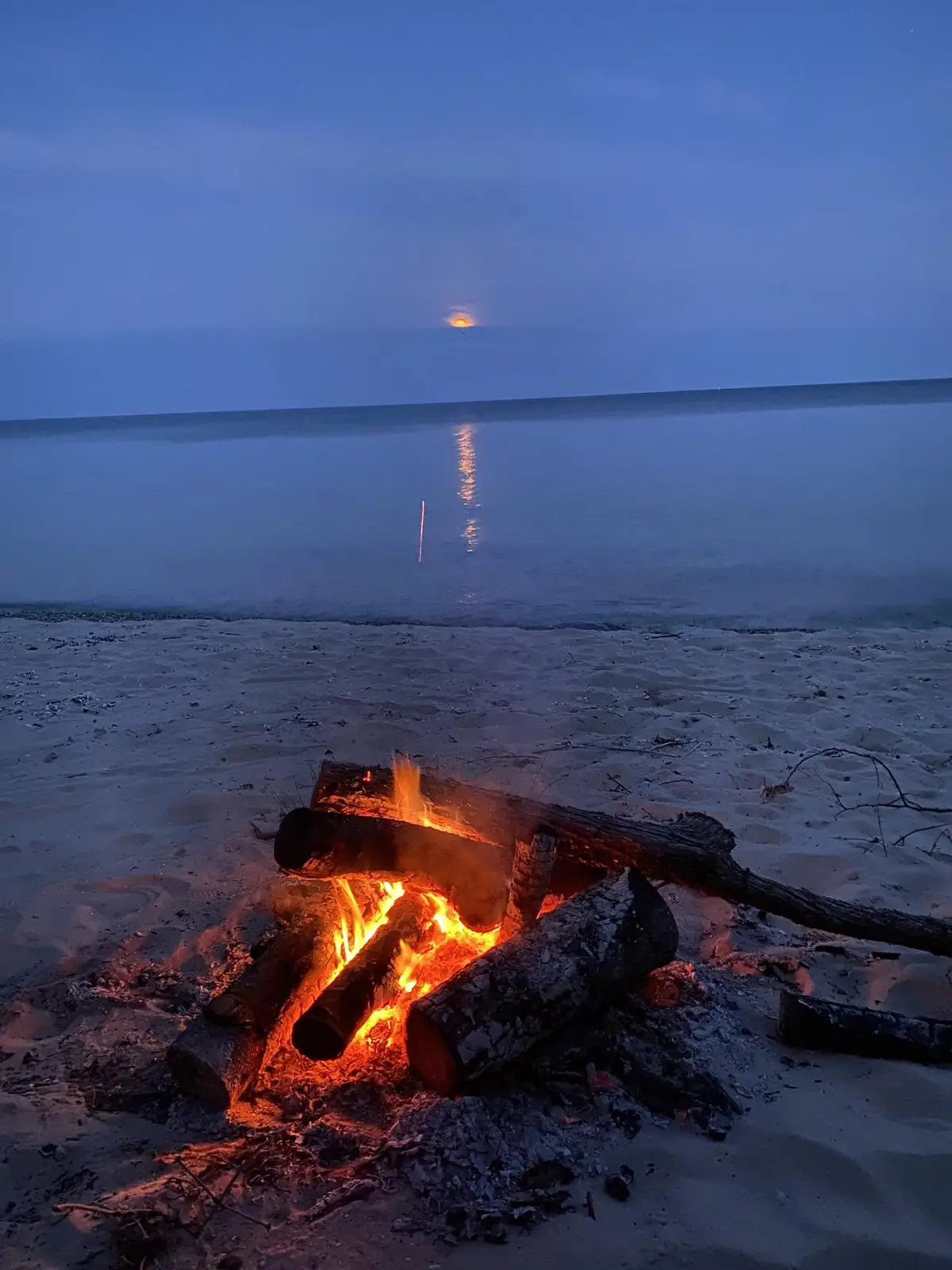 A campfire burns on a sandy shore beneath an orange moon reflected in calm water.