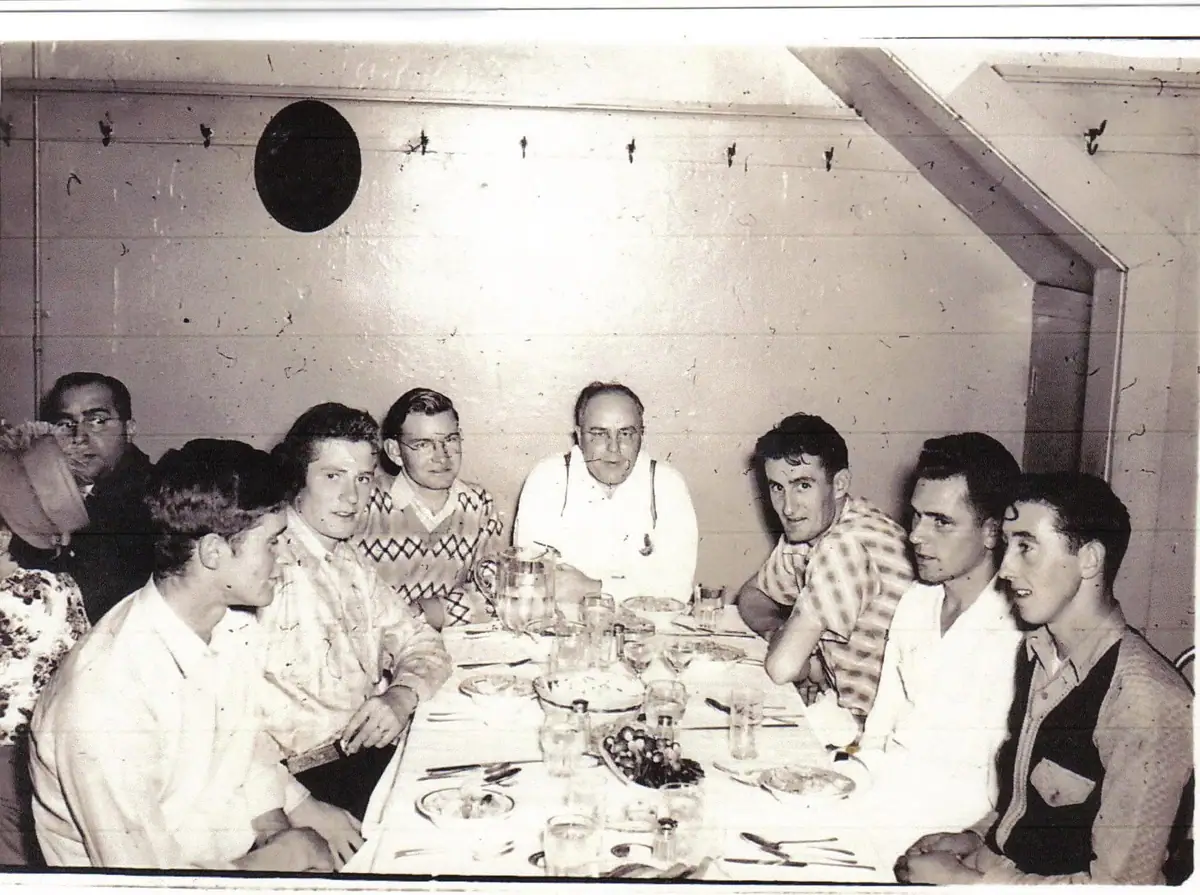A group of men gathers around a set dining table in a wood-paneled room.