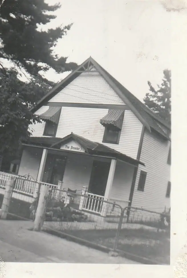 Black-and-white photograph of a two-story wood-sided house with a porch, striped awnings, and a fenced yard.
