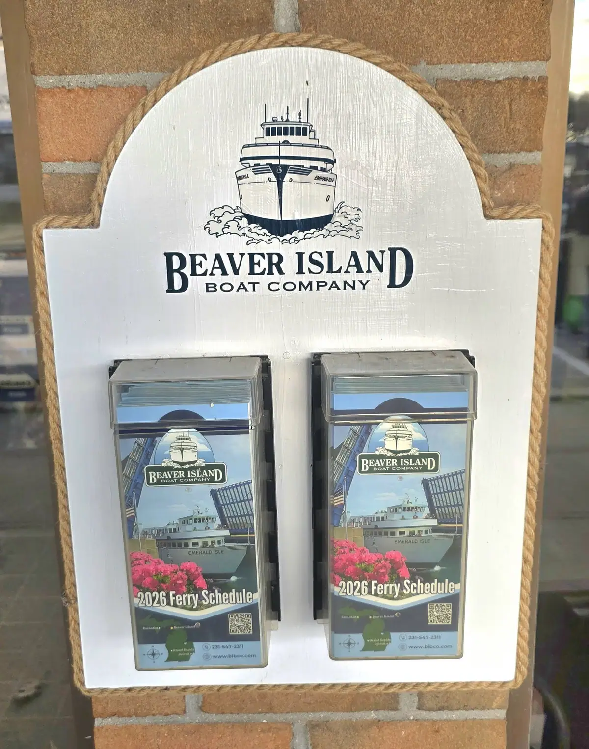 A rope-trimmed display plaque holds two ferry-schedule brochures against a brick wall.