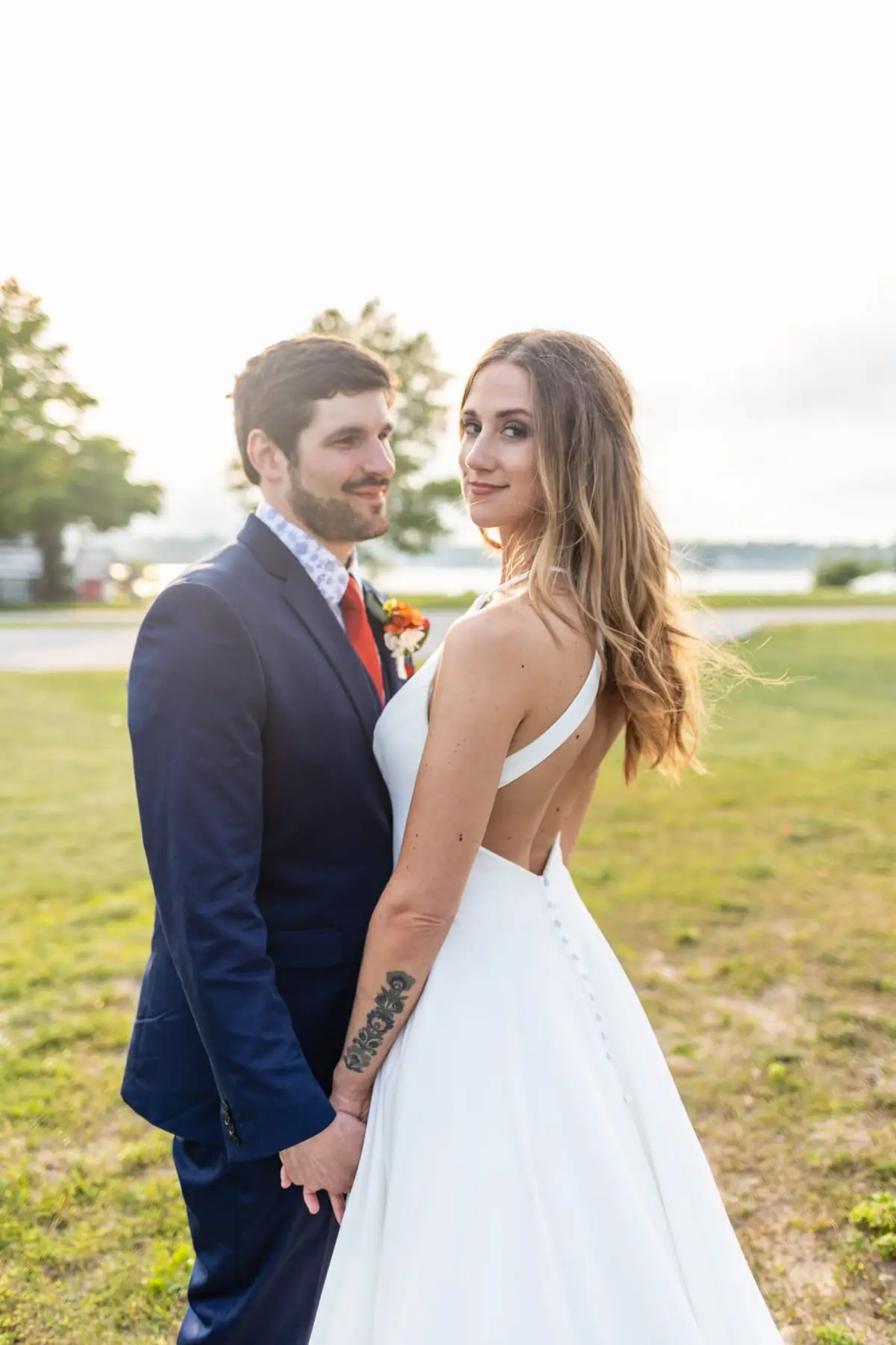 A couple in wedding attire stands hand in hand beside grassy grounds and water.