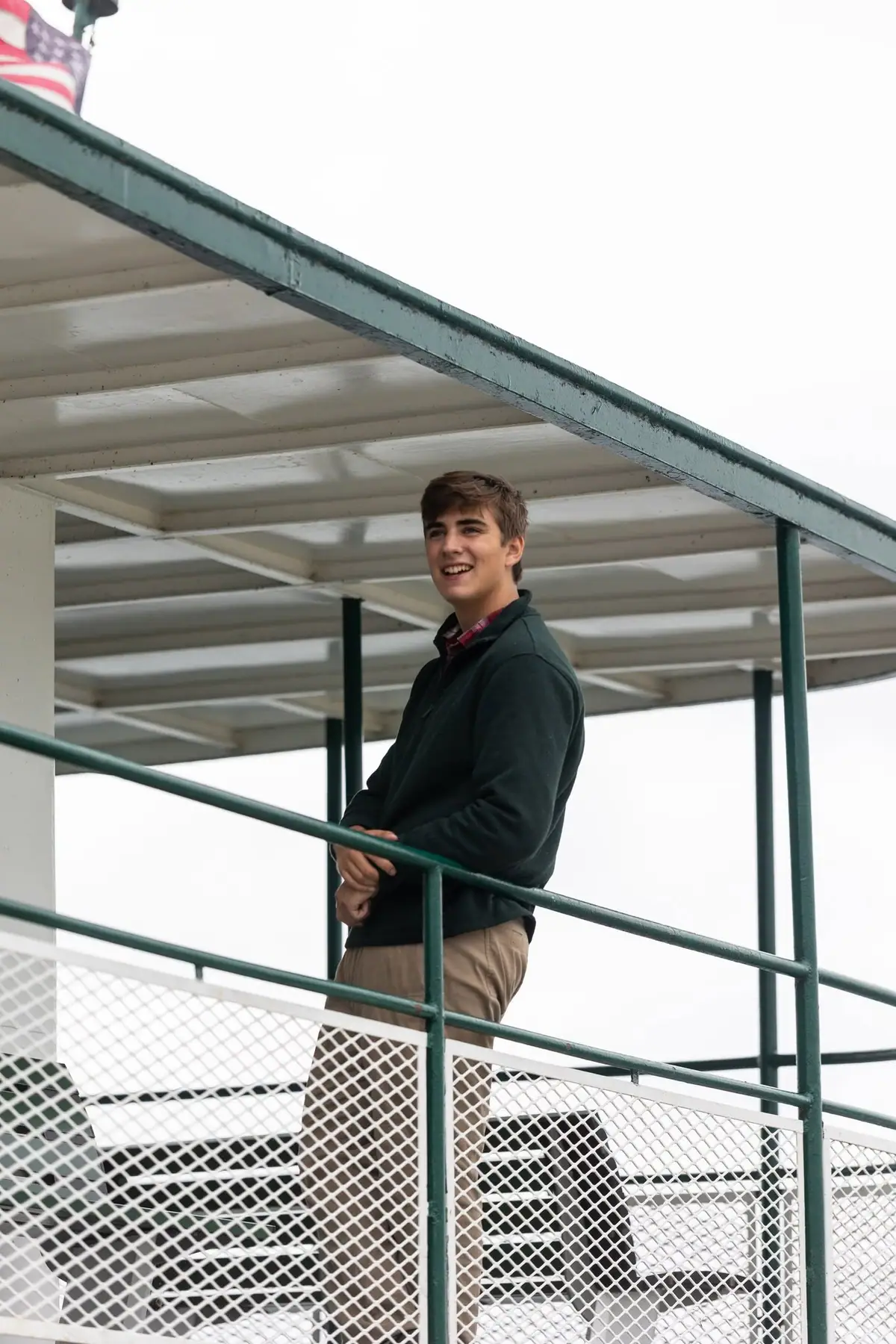 Smiling young man leaning against a green railing on a covered metal spectator stand.