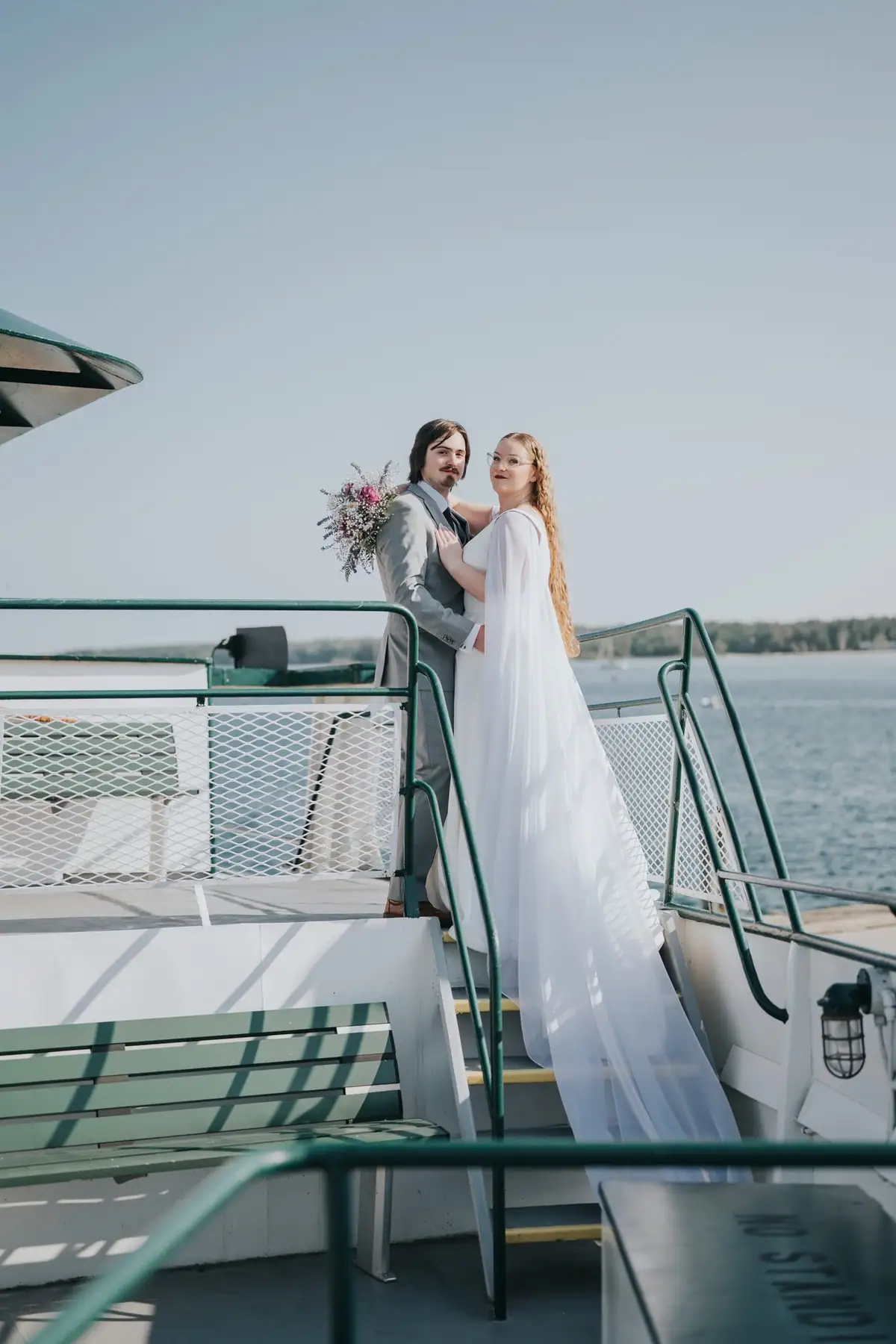 A newlywed couple poses on the deck of a passenger vessel beside green railings and benches.
