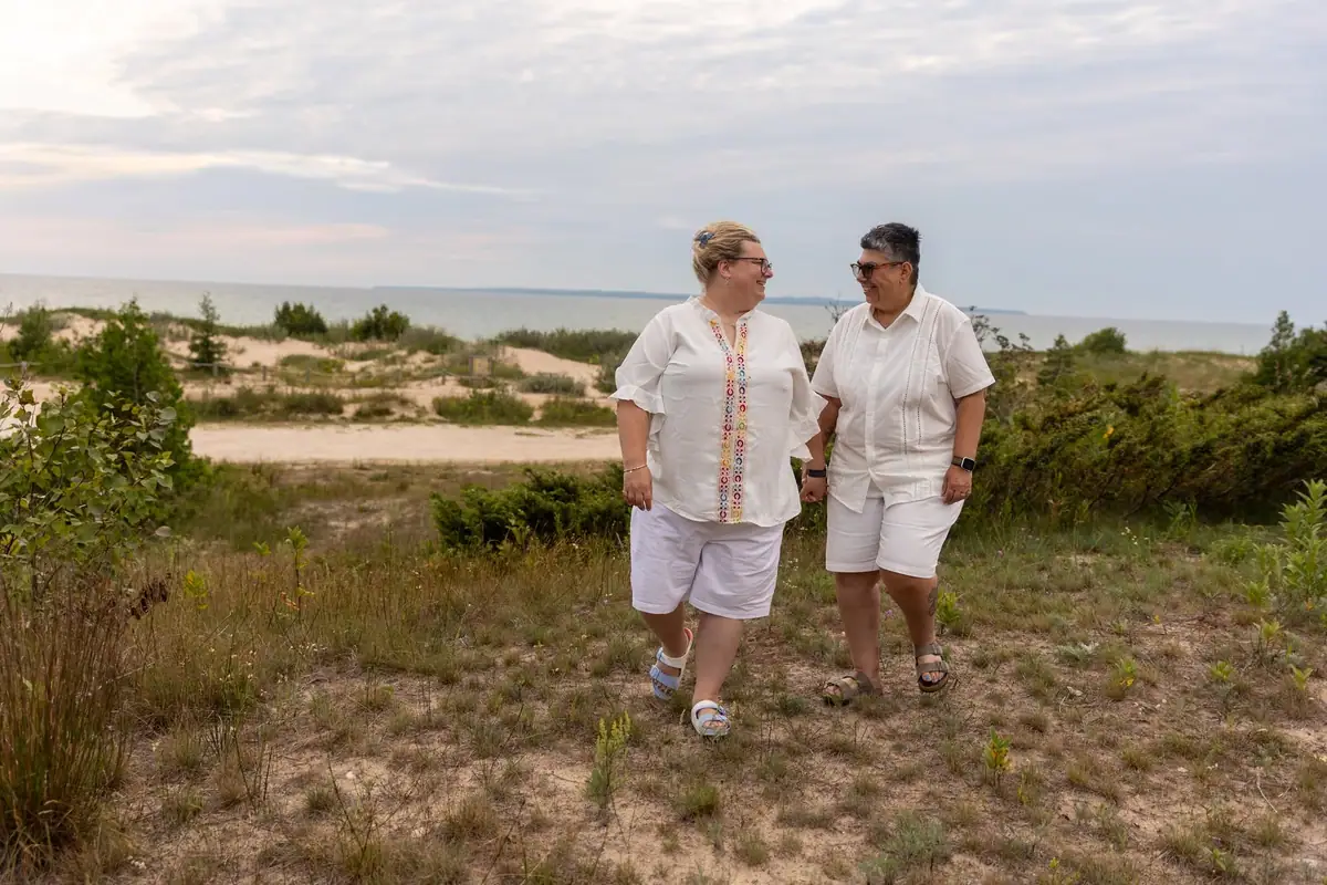 Two adults walk through sandy dune vegetation beside open water.