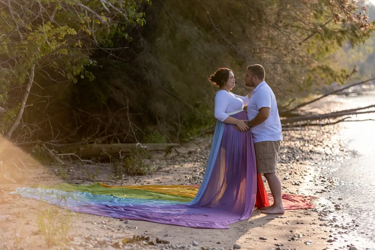 A couple poses beside a pebbled shoreline with rainbow-colored fabric trailing across the beach.