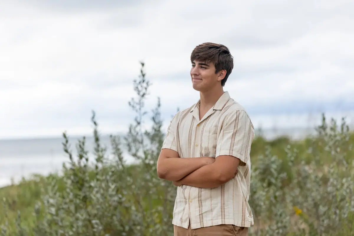 Portrait of a young man standing with folded arms among shrubs beside a body of water under a cloudy sky.