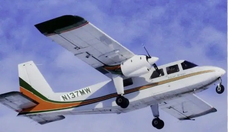 A white twin-engine propeller aircraft flies against a blue and cloudy sky.