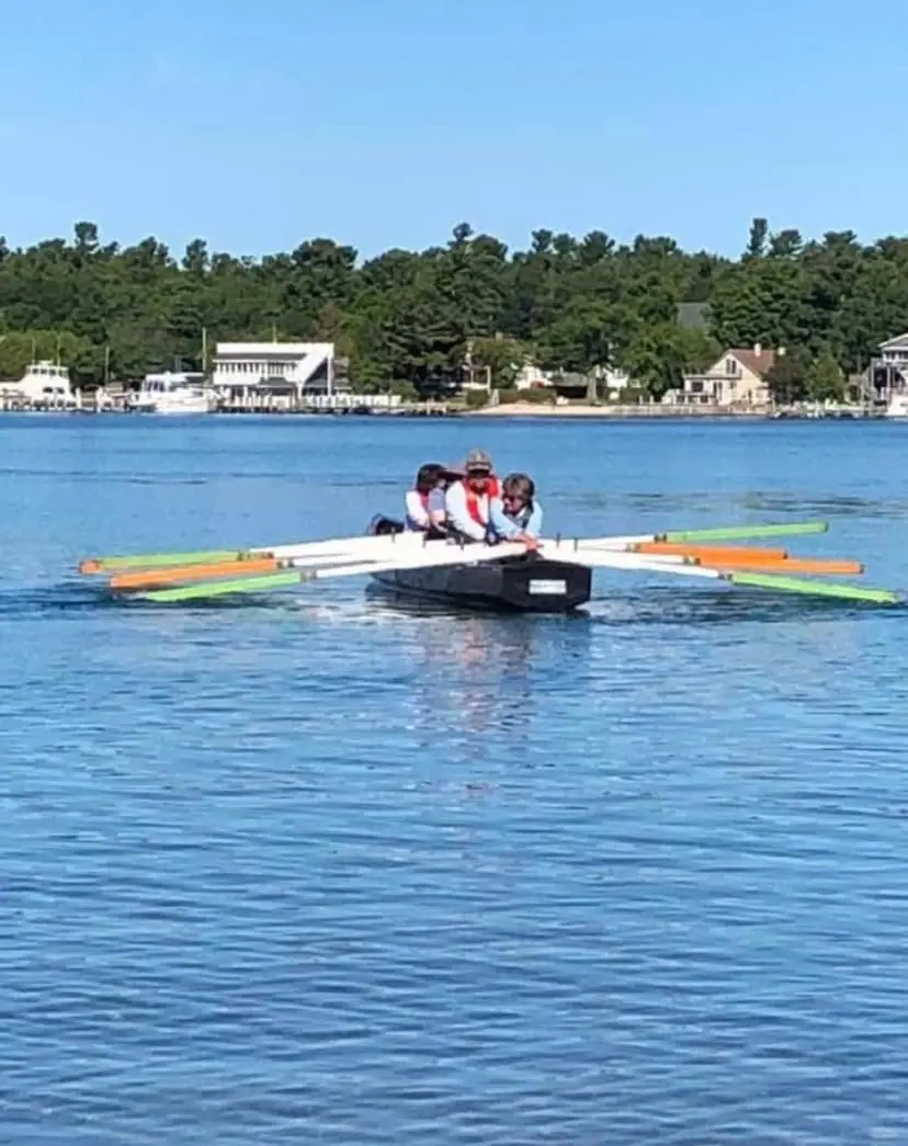 Four people in a black rowing shell cross calm blue water near a wooded shoreline and waterfront buildings.