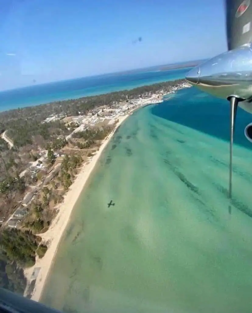 Aerial view of a sandy shoreline, wooded settlement, turquoise water, and a small aircraft wing and strut.