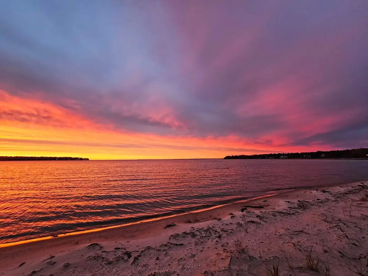 A vivid sunset spreads pink, orange, and purple light over rippled water and a sandy shoreline.