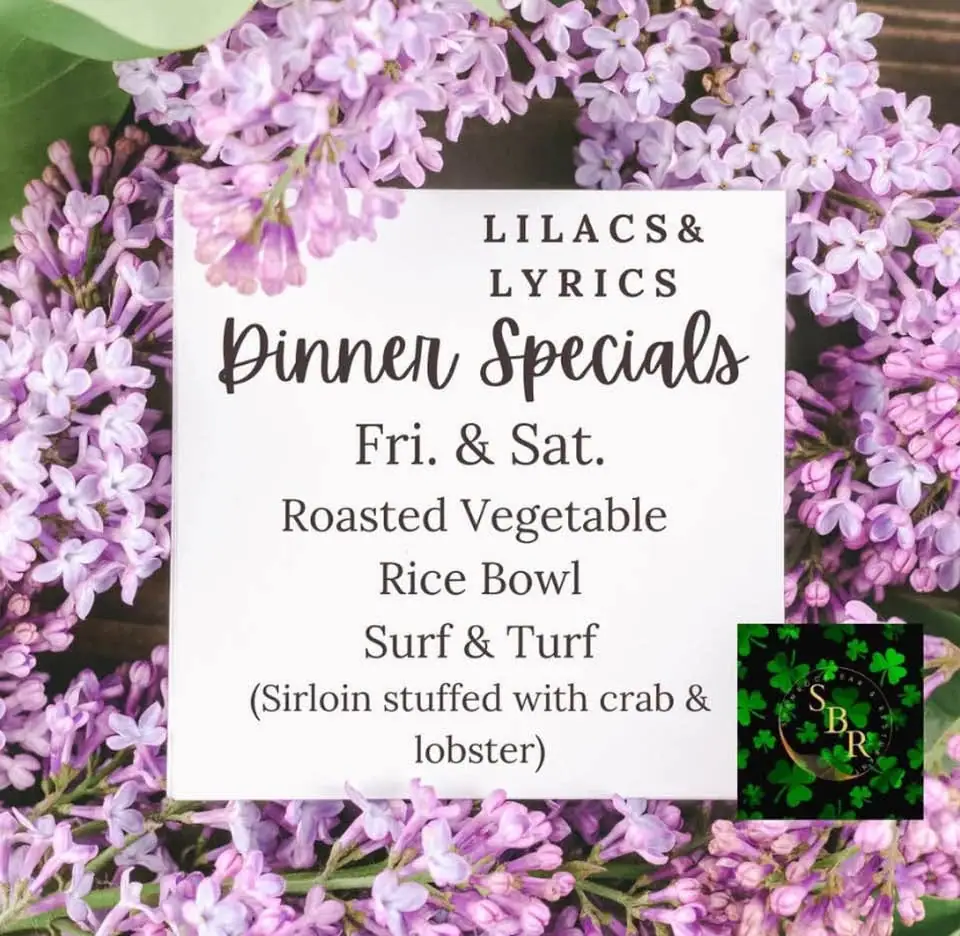 A printed dinner-specials sign surrounded by purple lilac blossoms, with a small shamrock logo in the lower corner.