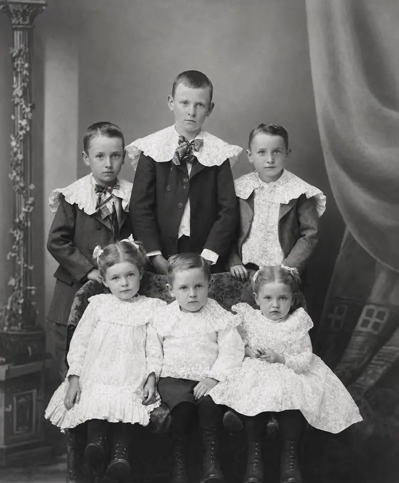 Studio portrait of six children in formal clothing, posed around an upholstered chair.