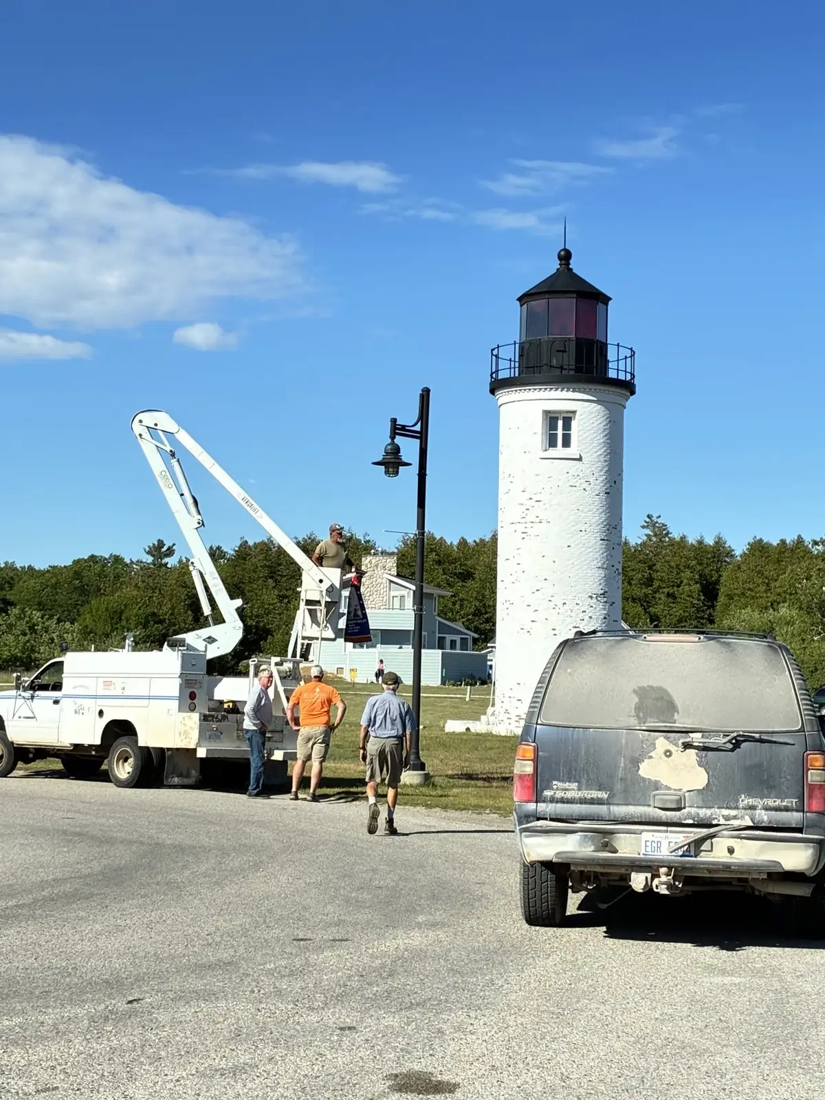 Workers use a boom lift beside a white lighthouse while vehicles stand along the paved roadside.