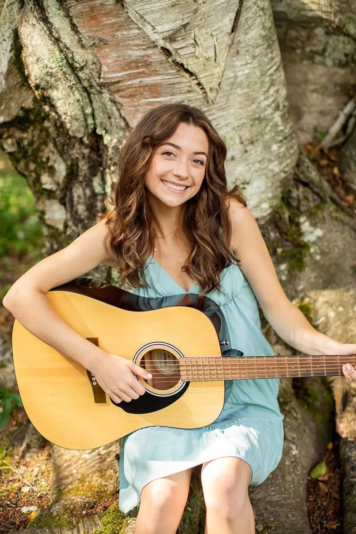 A smiling woman in a light blue dress holds an acoustic guitar in front of a large tree trunk.