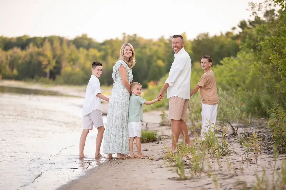 Five people hold hands while standing at the edge of shallow water on a sandy shoreline.