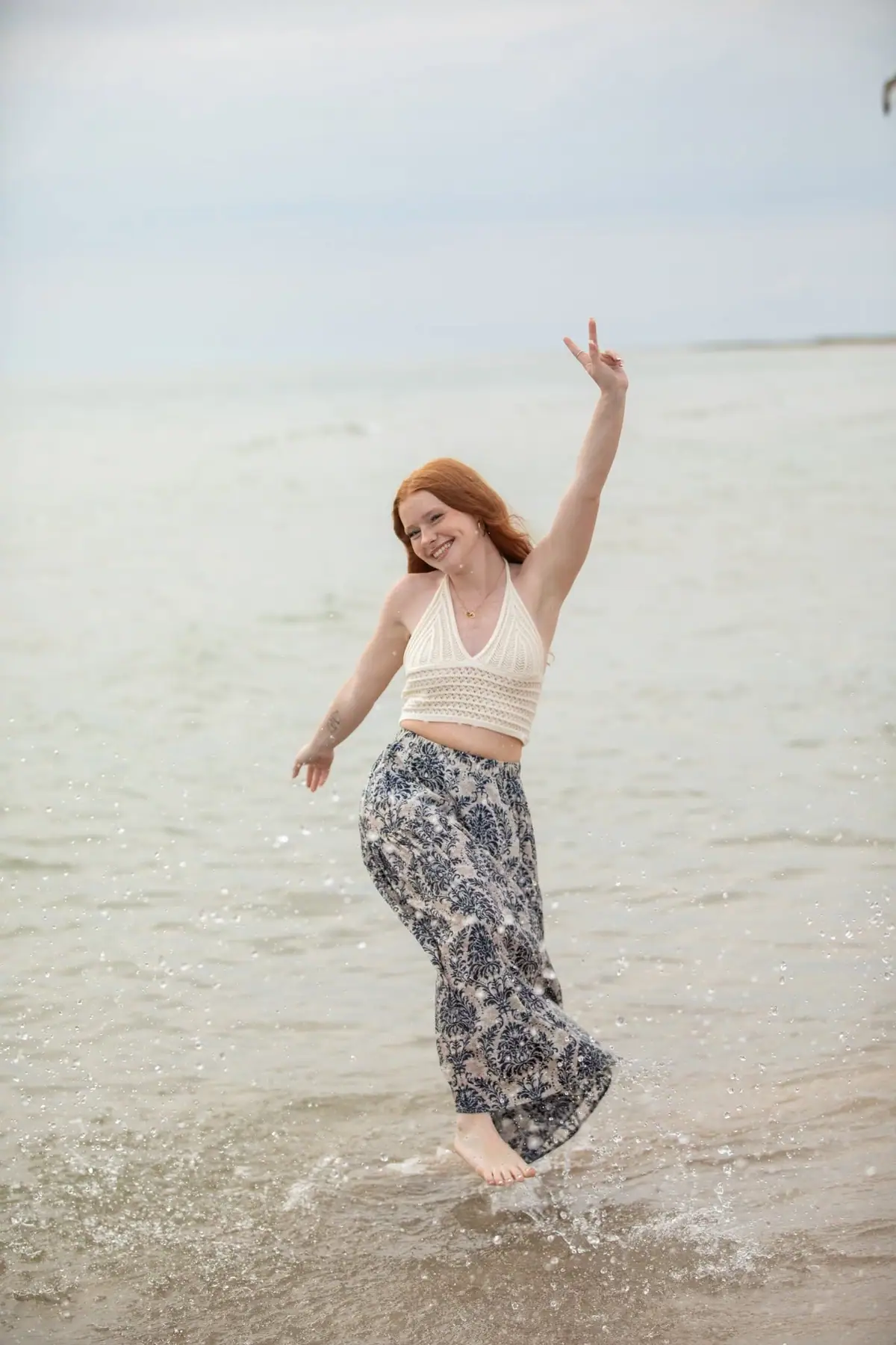 A red-haired woman smiles and raises a peace sign while jumping in shallow water near shore.