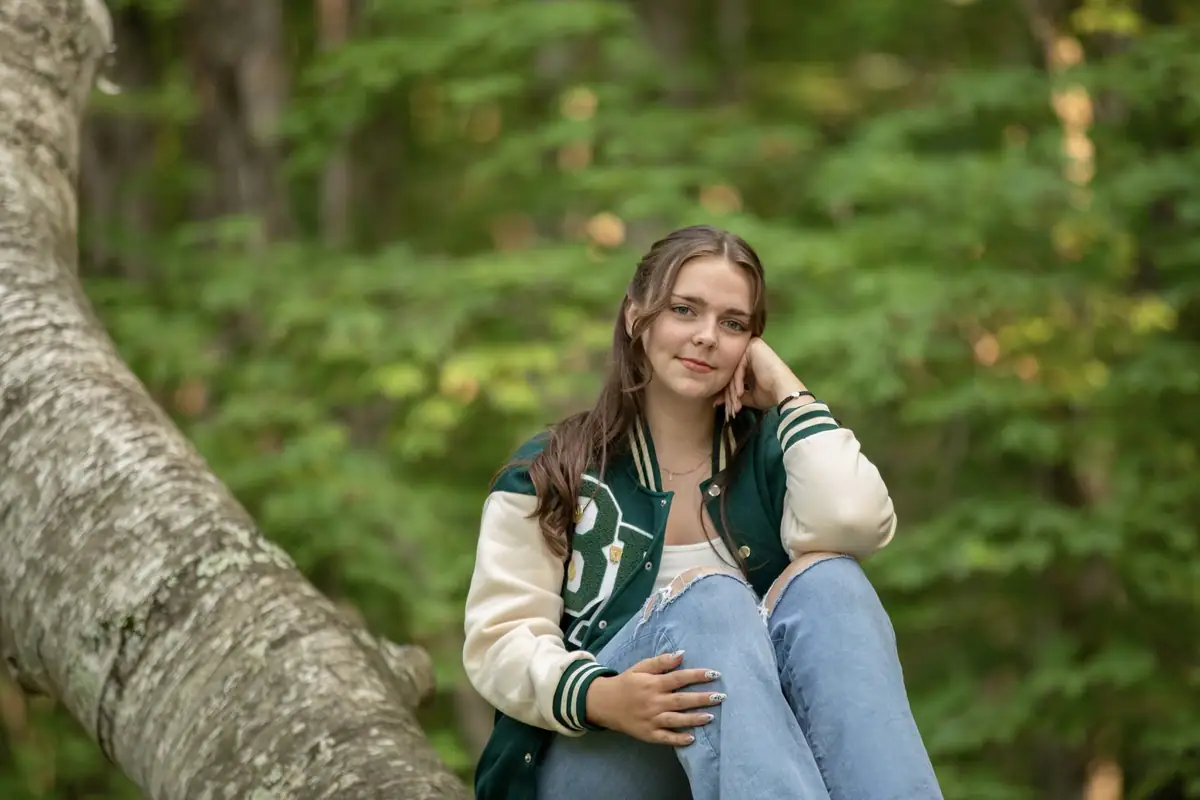A woman in a varsity jacket sits on a large tree branch in a green forest.