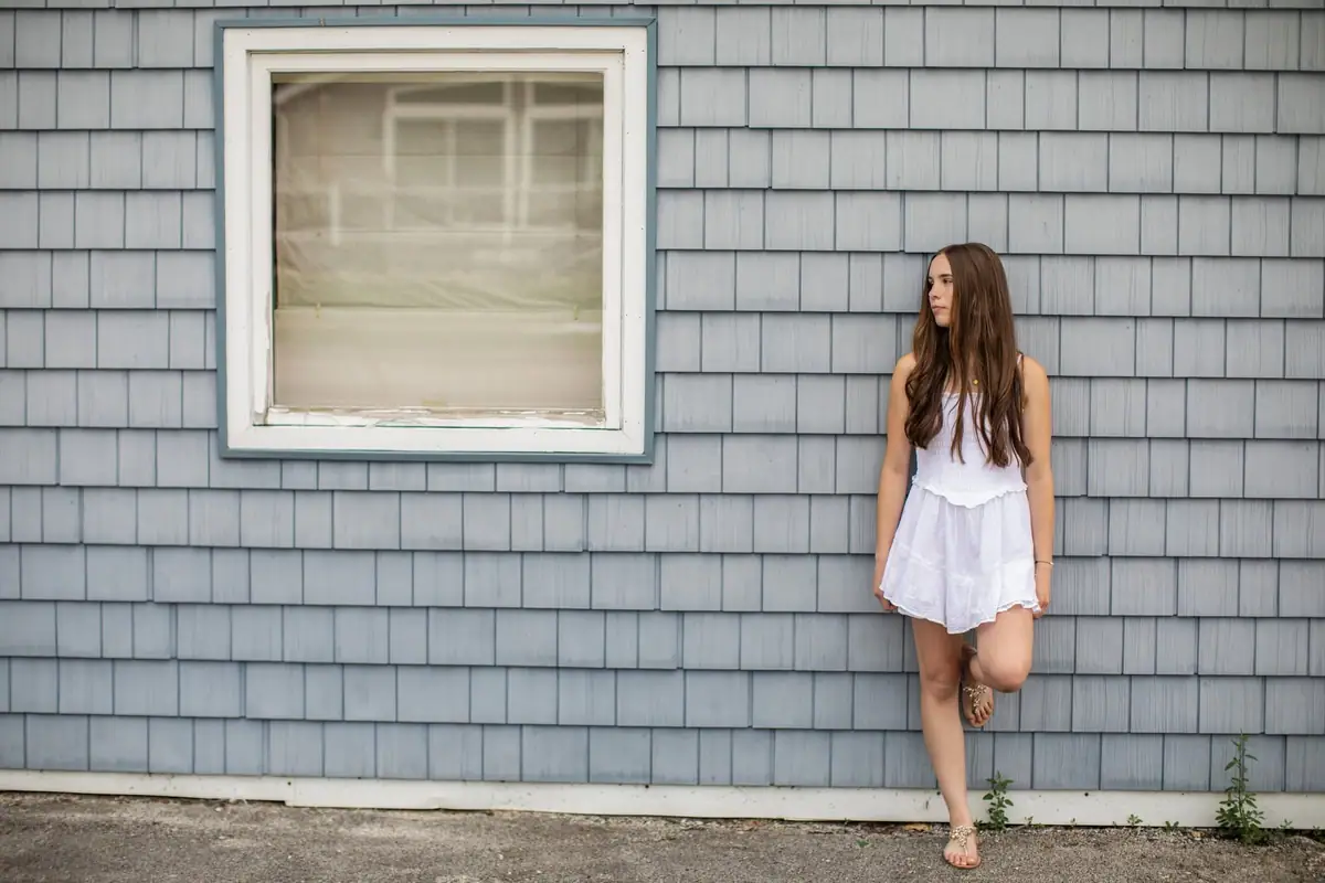 A young woman in a white dress poses beside a window on a blue-shingled exterior wall.