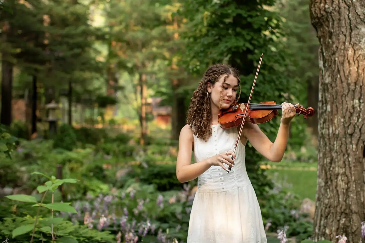 A young woman plays a violin beside a tree in a wooded garden.