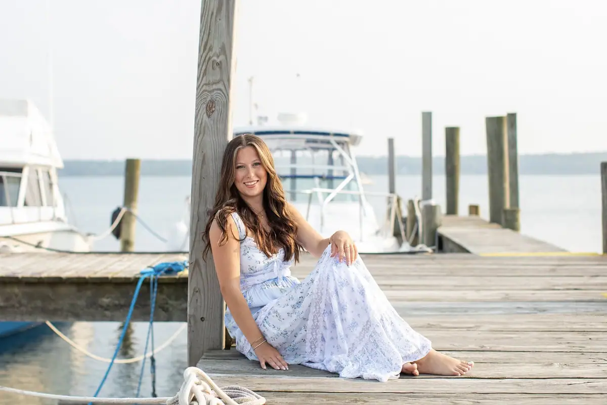 A smiling woman in a floral dress sits barefoot on a wooden dock beside moored boats.