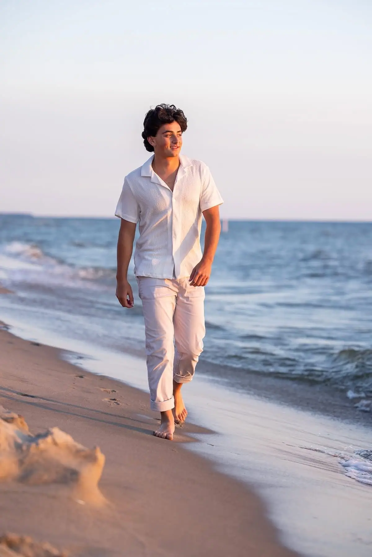 A young man walks barefoot along a sandy shoreline in warm evening light.