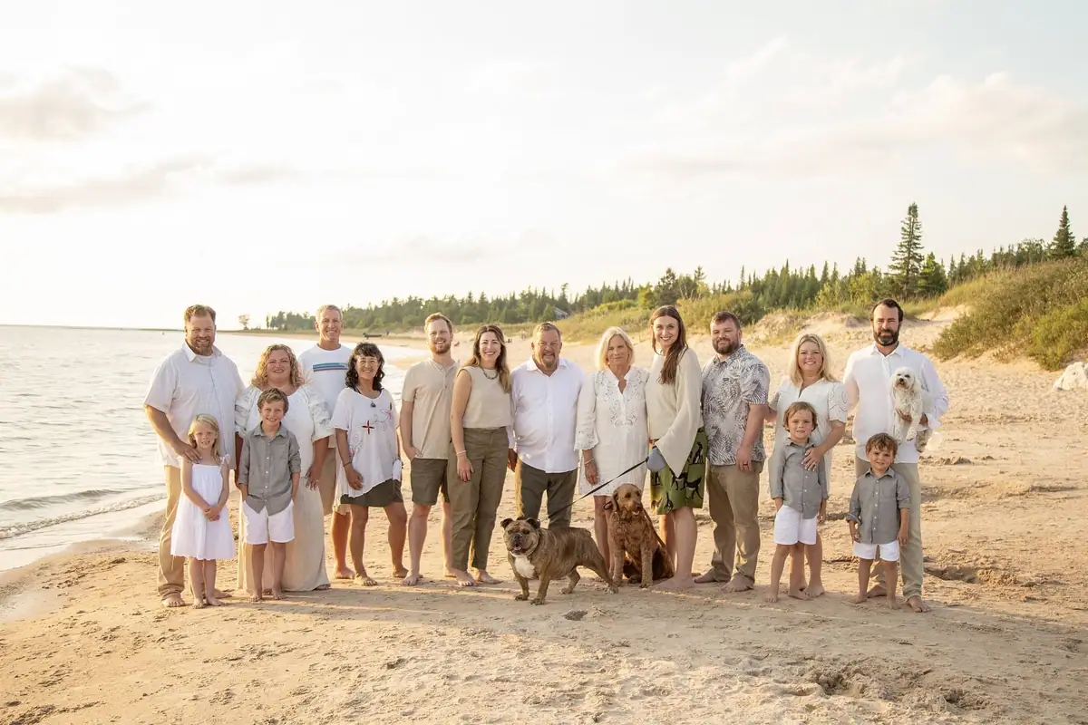 A large group poses with several dogs on a sandy shoreline beside wooded dunes.