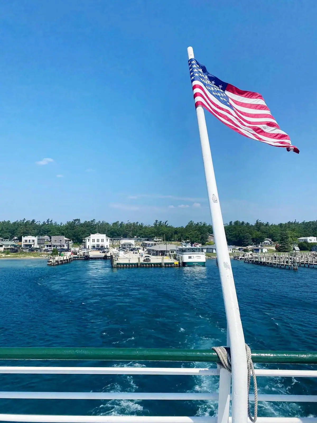 American flag flying from a boat above a waterfront harbor with docks and a ferry.