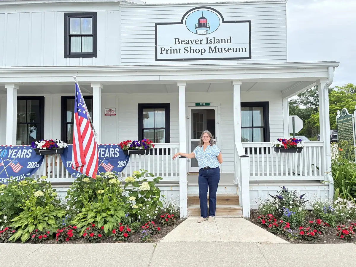 A woman stands in front of a white museum building with a lighthouse sign, porch, flag, and flower gardens.