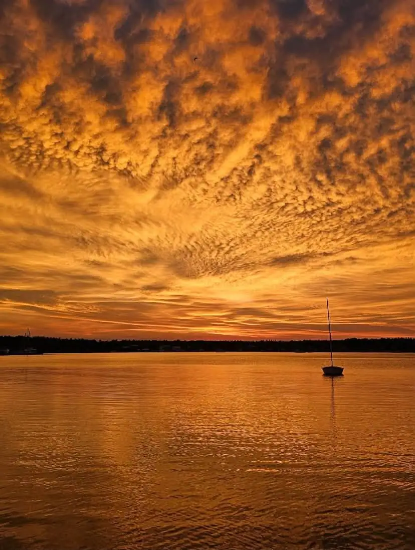 A small sailboat sits on calm water beneath a richly textured orange sunset sky.