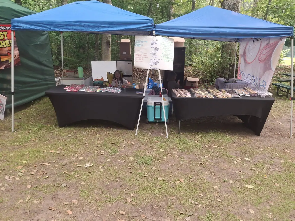 Outdoor vendor booths under blue canopies display packaged goods, a cooler, and a handwritten menu board.