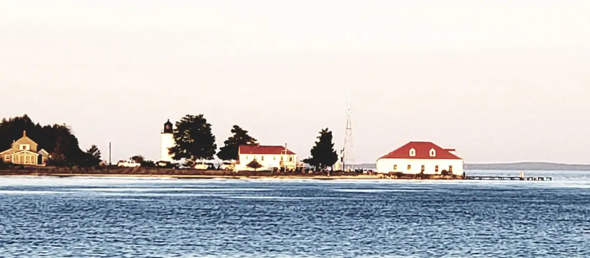 Waterfront view of a lighthouse, houses, trees, antenna tower, and pier along a narrow shoreline.