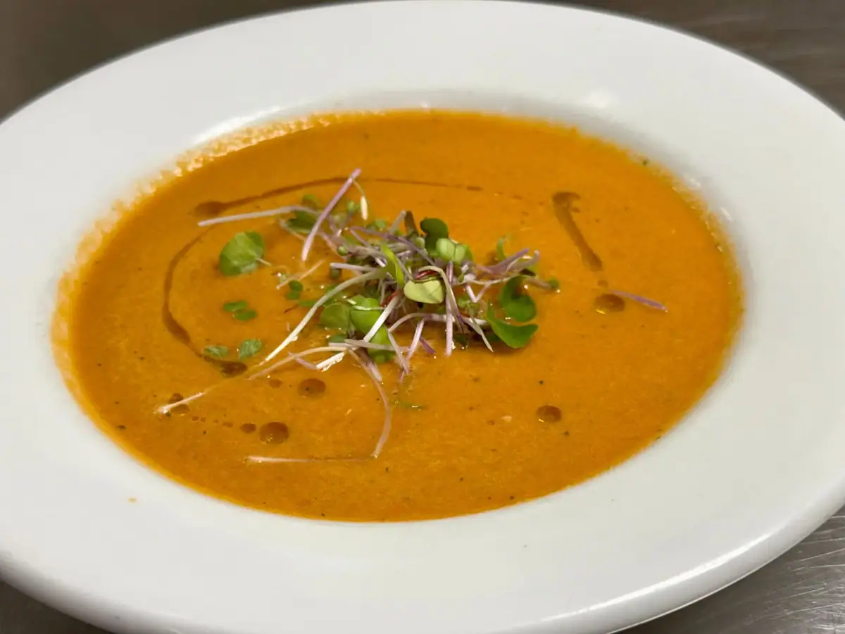 A white bowl holds orange soup garnished with green and purple microgreens.
