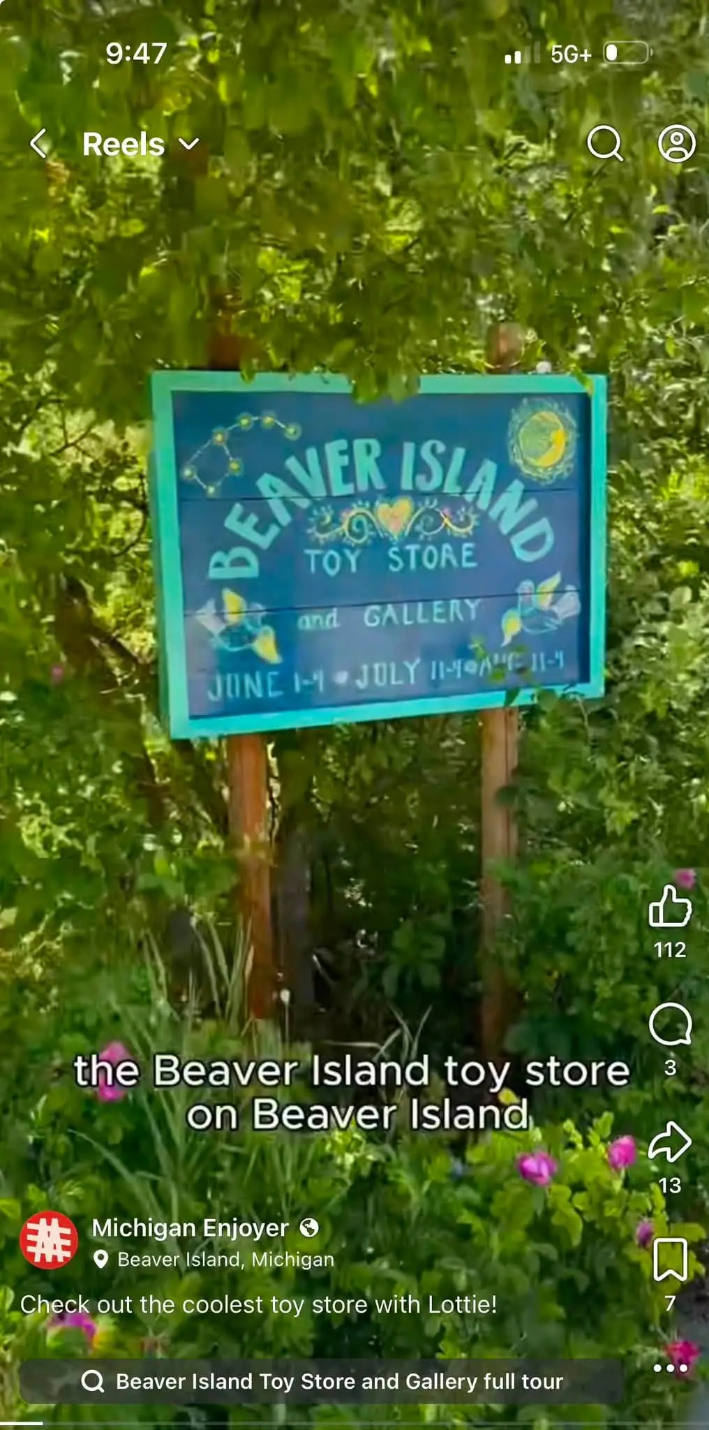 A painted blue sign for a toy store and gallery stands among leafy trees and flowers.