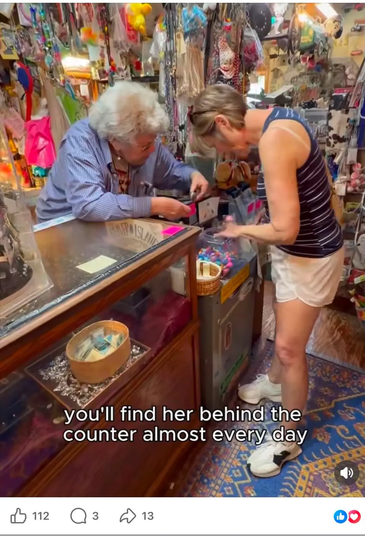 Two women interact across a glass display counter in a crowded shop filled with toys and other merchandise.