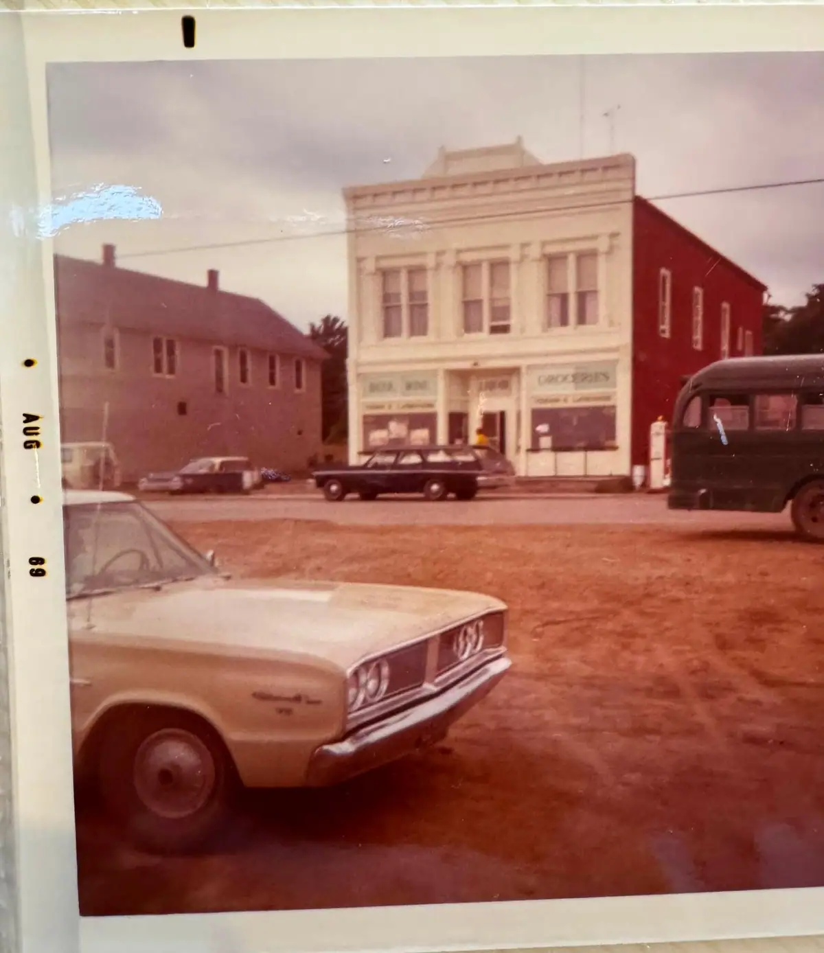 Color photograph of a storefront-lined street with vintage automobiles and a white-bordered print edge.