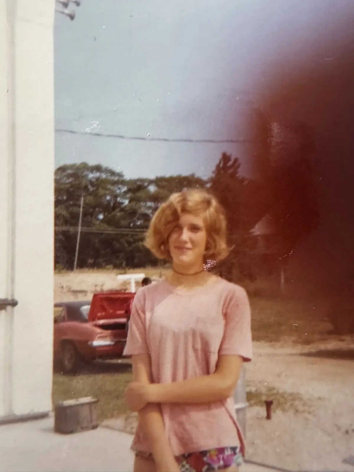 A young woman in a pink shirt and floral shorts stands outdoors beside a red car.