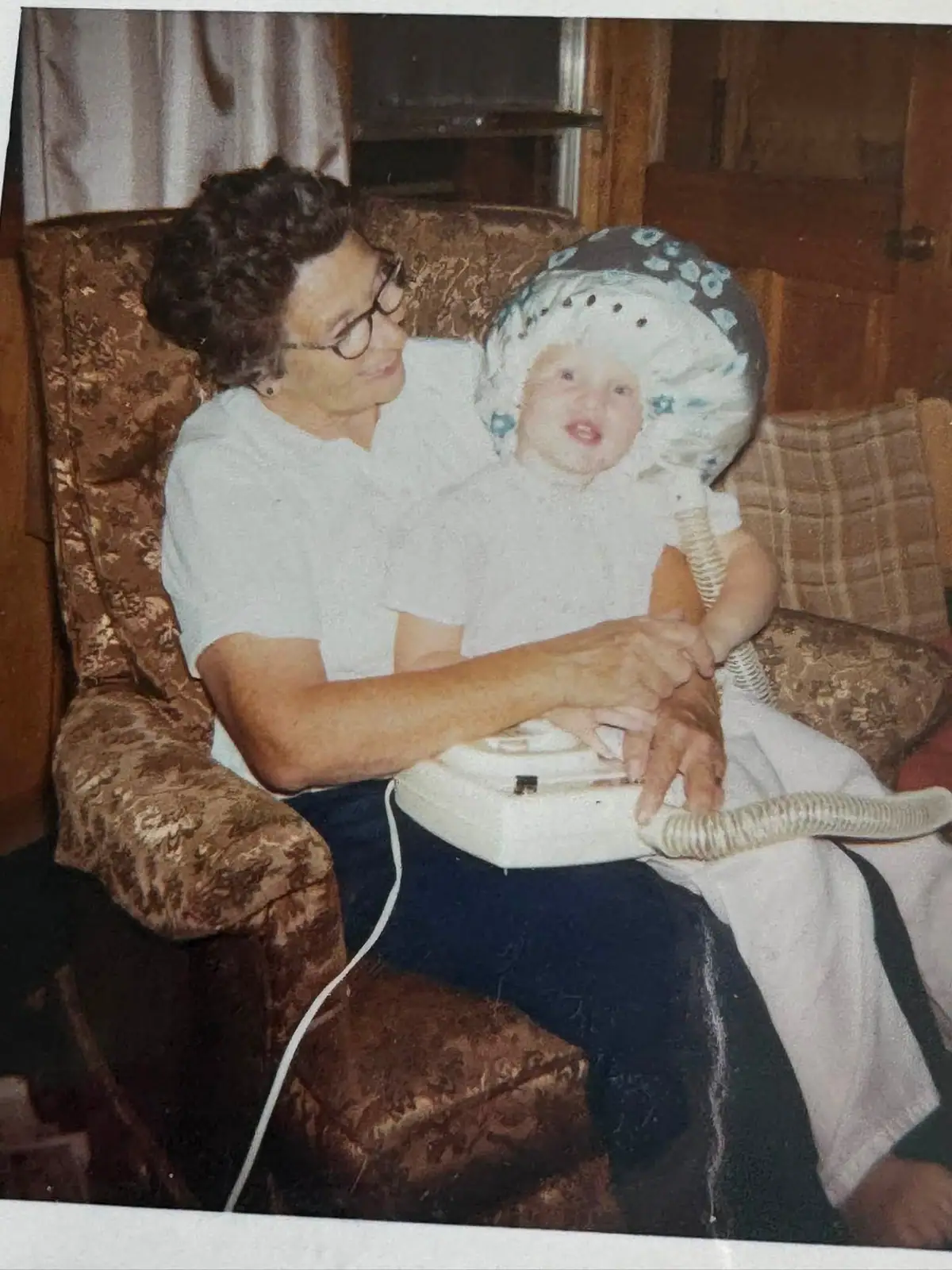 An older woman with glasses holds a smiling baby in a patterned armchair.