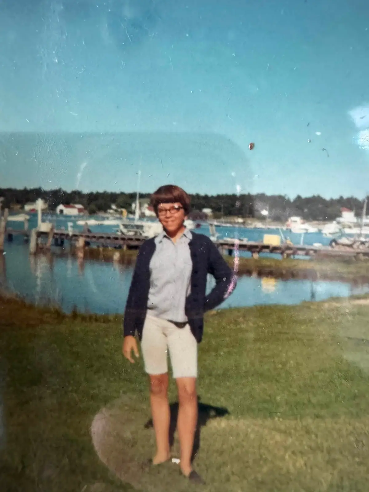 A person stands on a grassy bank beside a harbor with boats and a pier.
