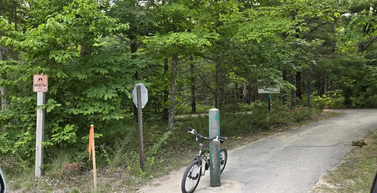 A bicycle rests against a post beside a paved path in a wooded area, with trail and traffic signs nearby.