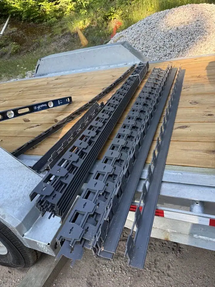 Black landscape edging and a spirit level rest on the wooden deck of a utility trailer beside a gravel pile.